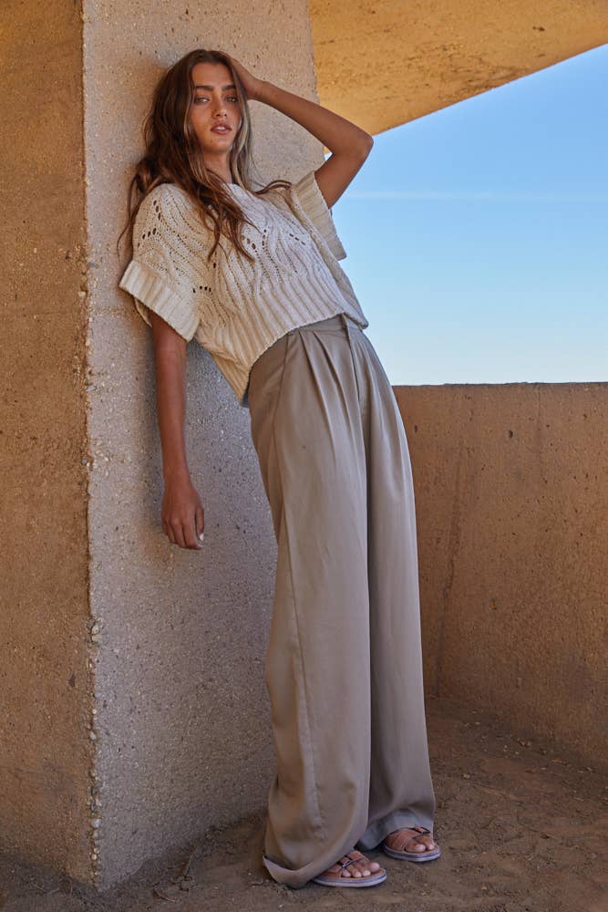 Woman in a light-colored top and beige pants standing against a textured wall with a clear sky.